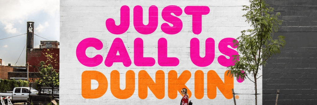 A photo of Dunkin's rebranding slogans painted on a brick wall. The text reads "Just call us Dunkin'" in sans-serif, rounded font.